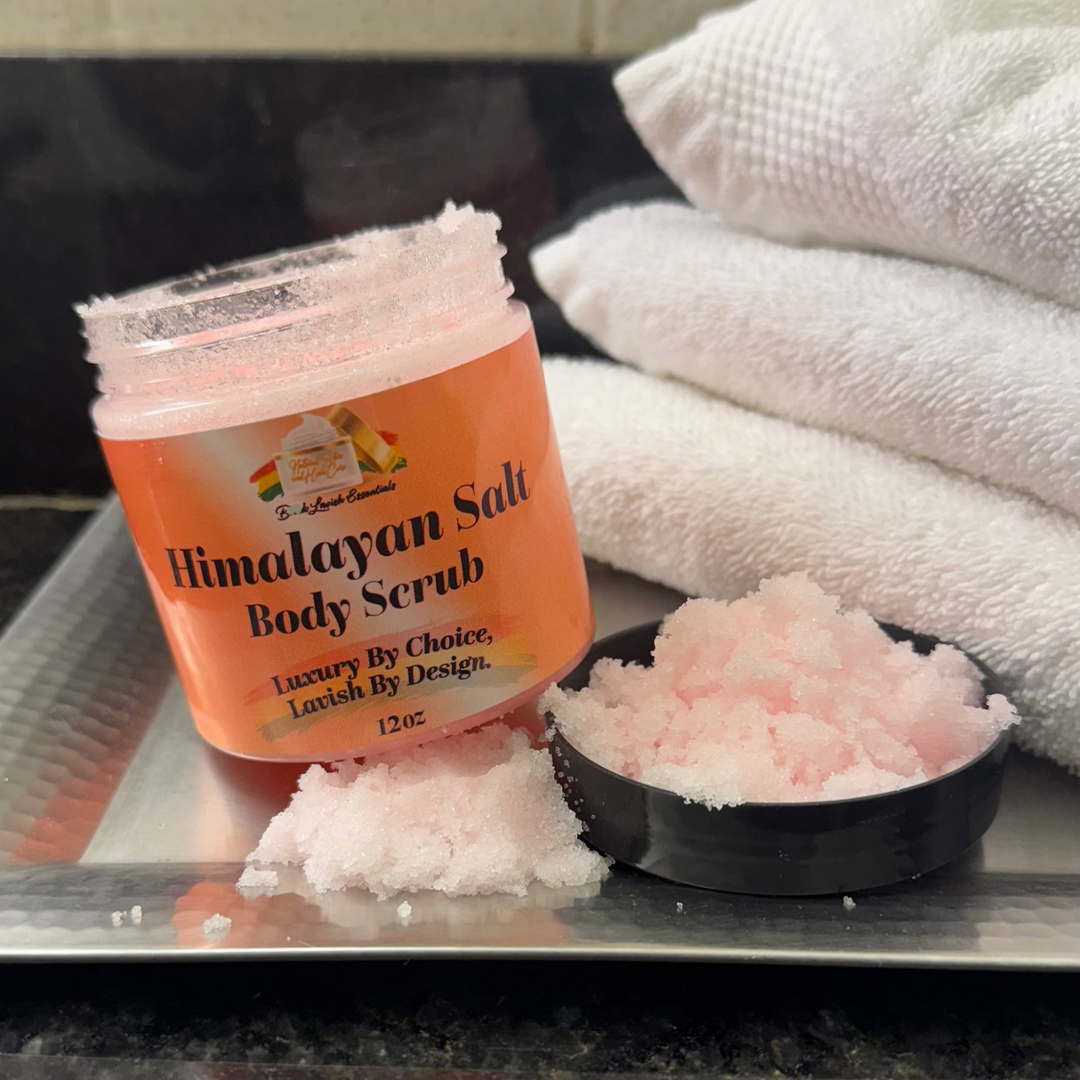 A jar of Himalayan Salt Exfoliating Body Scrub is open with some scrub visible outside the jar. The jar is labeled with the product name and has a pink hue, corresponding to the salt content.
