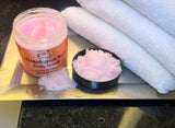 A jar of pink Himalayan salt body scrub is open with some product spilled out onto a surface next to a black dish and a white towel.